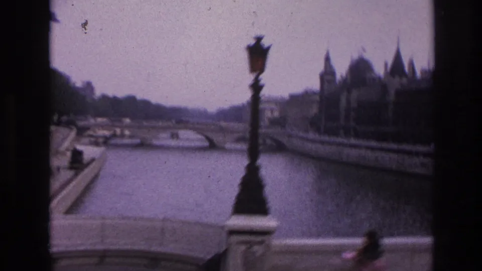1967 Paris home-movie frame showing a riverside bridge, ornate streetlamp, stone quay, and large historic buildings across the water.