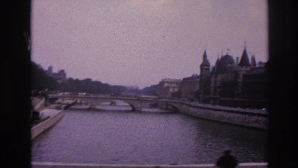 1967 Paris river view with arched bridge spans, stone embankments, historic riverside buildings, and a blurred pedestrian at the bottom edge.