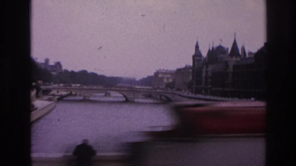 1967 Paris home-movie frame showing the Seine, a low bridge, historic riverside buildings, and a dark vehicle or window edge in the foreground.