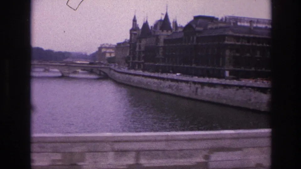 1967 Paris home-movie frame showing the Seine embankment, an arched bridge, and a large historic riverside building with spires and steep rooflines.