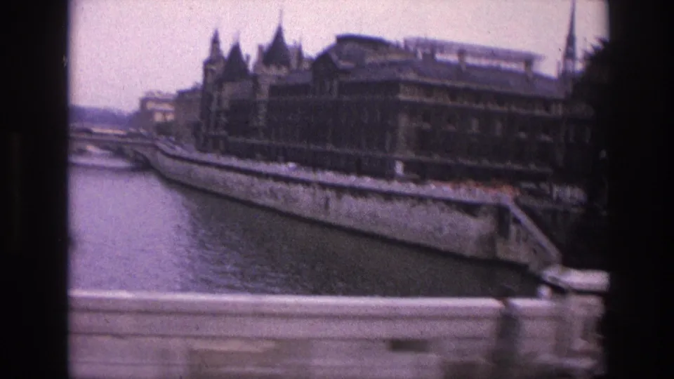 1967 Paris frame with the Seine, stone embankment, ornate riverside building, pale bridge railing, and dark vehicle edge in the foreground.