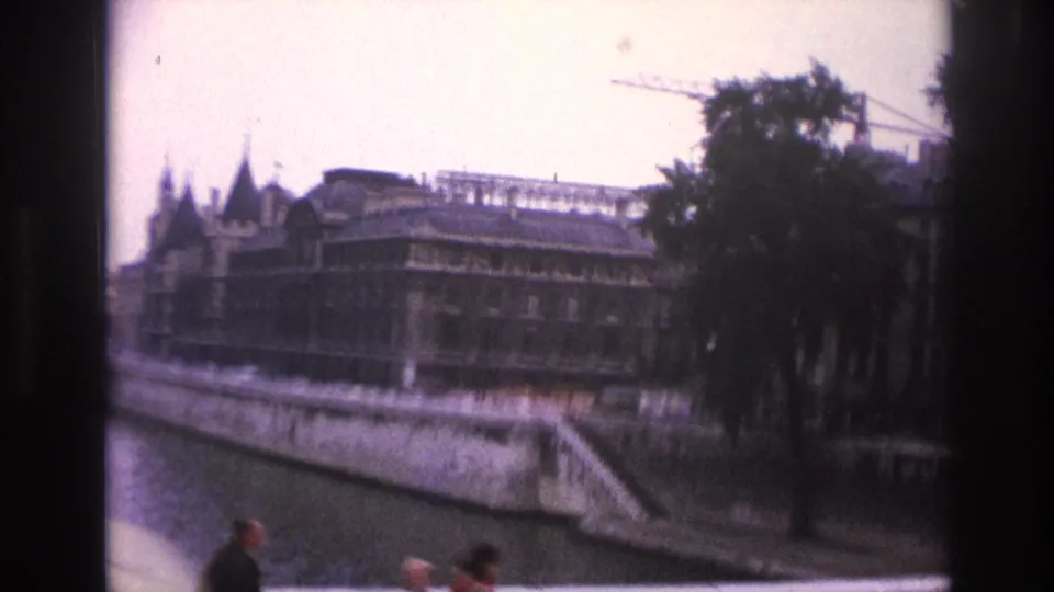 Paris, 1967, viewed from a moving car along the Seine, with riverside buildings, a stone quay, trees, a crane, and small pedestrians.