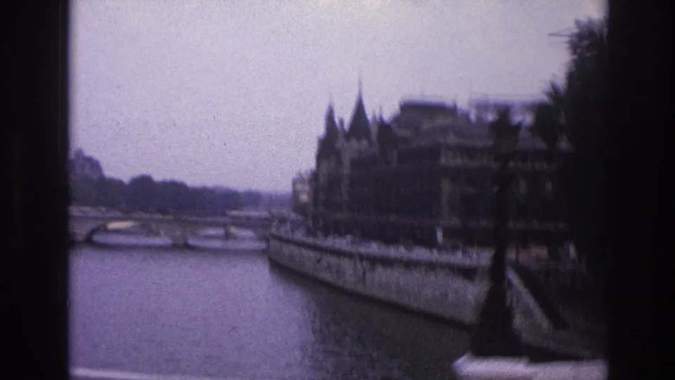 Wider 1967 Paris view down the Seine with moored boats, a stone embankment, ornate riverside buildings with spires, and dark trees.