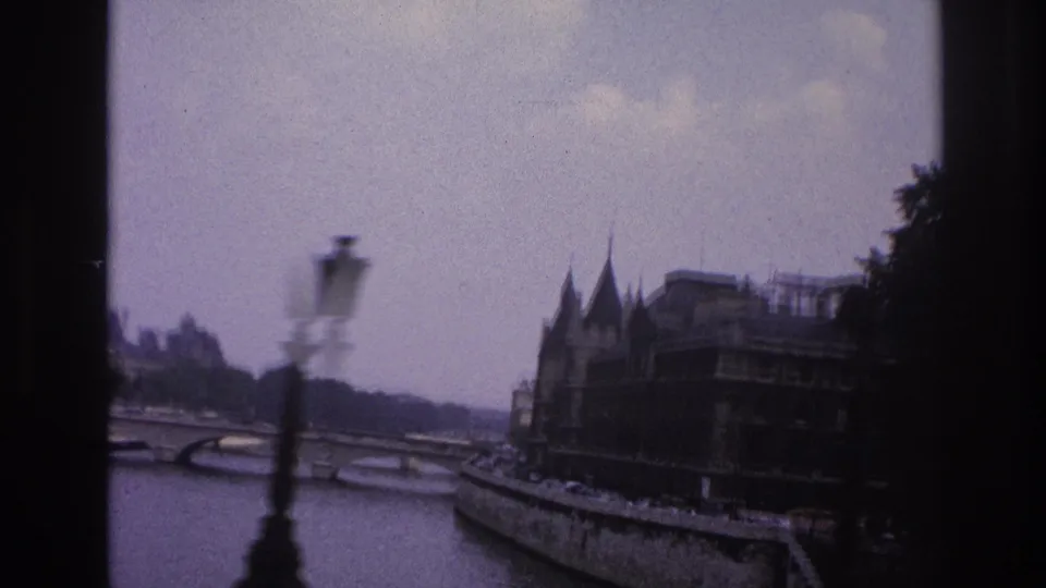 1967 Paris home-movie frame showing the Seine, a stone bridge, foreground lamppost, trees, and historic riverside architecture.