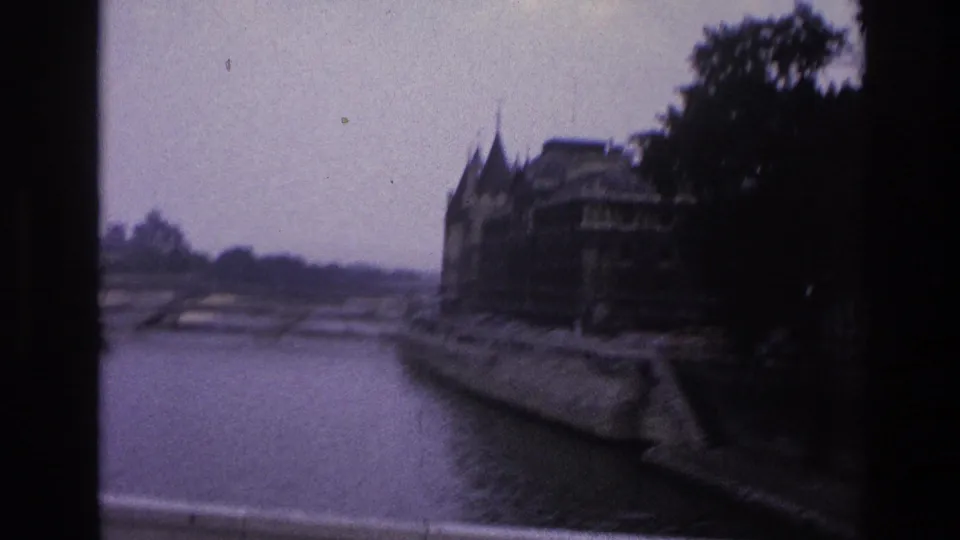 1967 Paris 8mm frame looking along the Seine toward bridge arches, quayside walls, trees, and older riverside buildings.