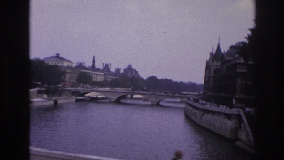 1967 Paris home-movie frame showing the Seine, bridge arches, distant civic-style architecture, trees, and quayside buildings.