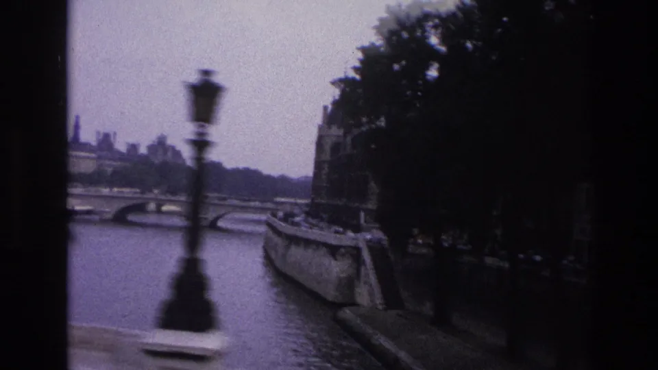 1967 Paris 8mm frame with a blurred lamppost passing before the Seine, stone bridge arches, quayside wall, and shaded trees.