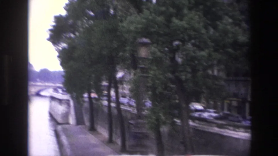 1967 Paris view along the Seine, with tree-lined quay, bridge spans, street lamps, and parked cars visible through soft home-movie focus.