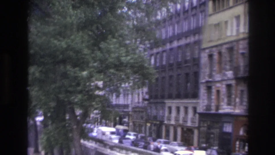 1967 Paris street scene with leafy foreground trees, tall residential or commercial buildings, sidewalks, and rows of period parked cars.