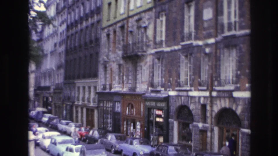 1967 Paris street view with rows of parked cars, older building facades, shopfronts, and small pedestrians visible near the sidewalk.