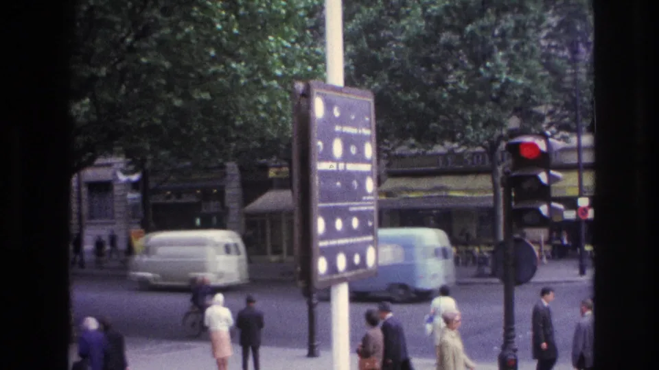 1967 Paris intersection with pedestrians crossing or waiting, a red traffic light, a street information sign, and a light van passing.