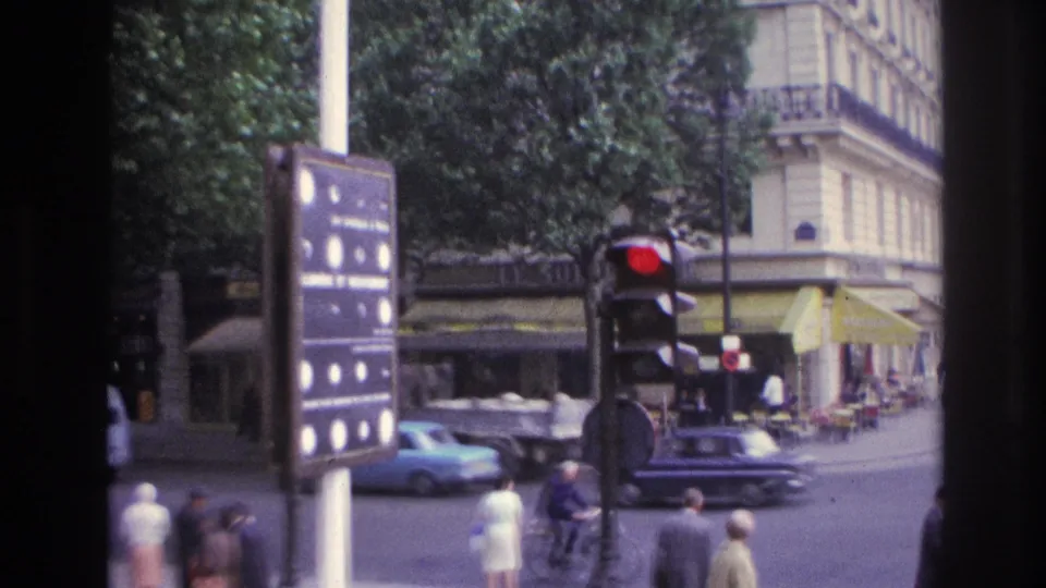 1967 Paris street corner showing pedestrians, cars, a red traffic light, shop awnings, trees, and a stone corner building.