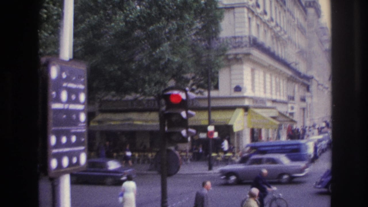 1967 Paris street corner with a red traffic light, pedestrians, vintage automobiles, café awnings, and pale stone architecture.