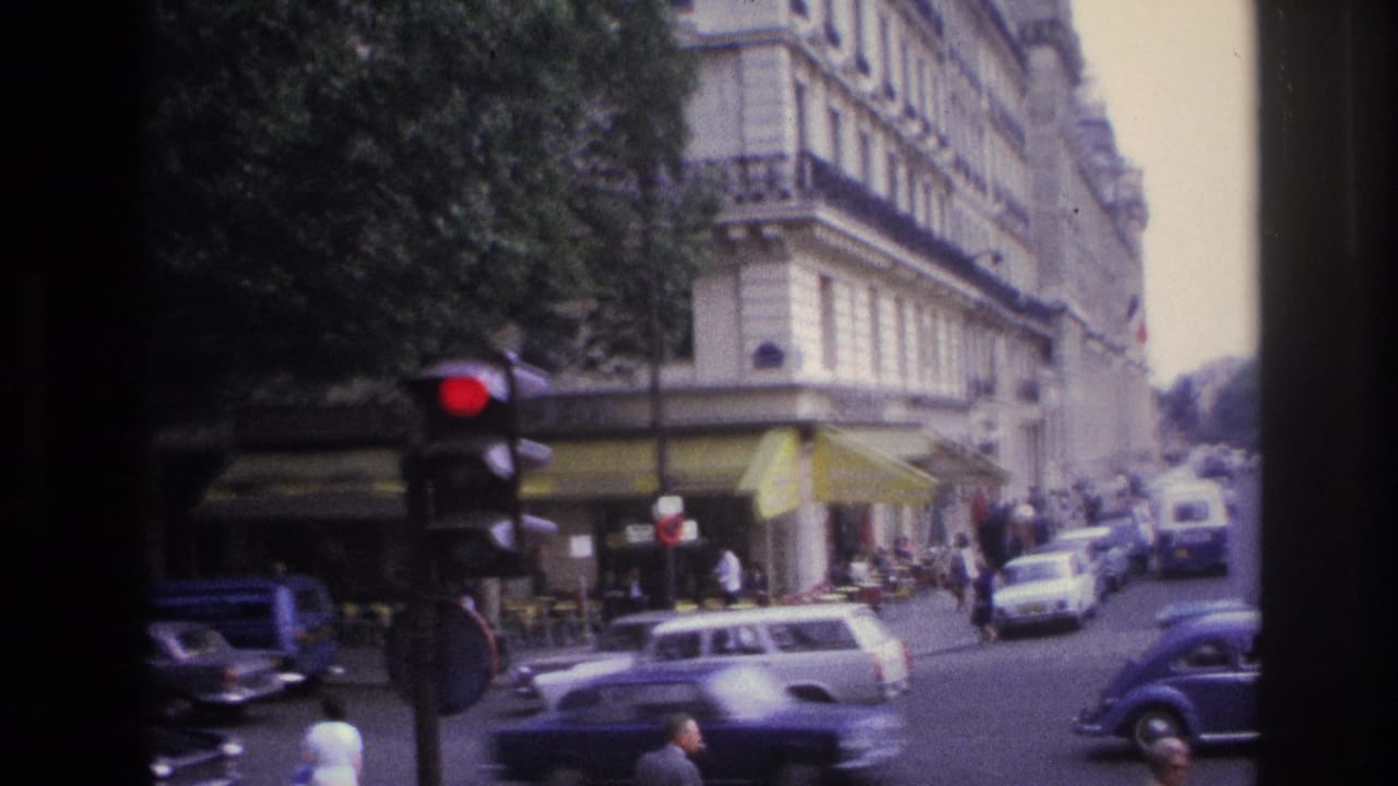 1967 Paris intersection with cars moving past a corner café, yellow awnings, pedestrians, and a red traffic signal.