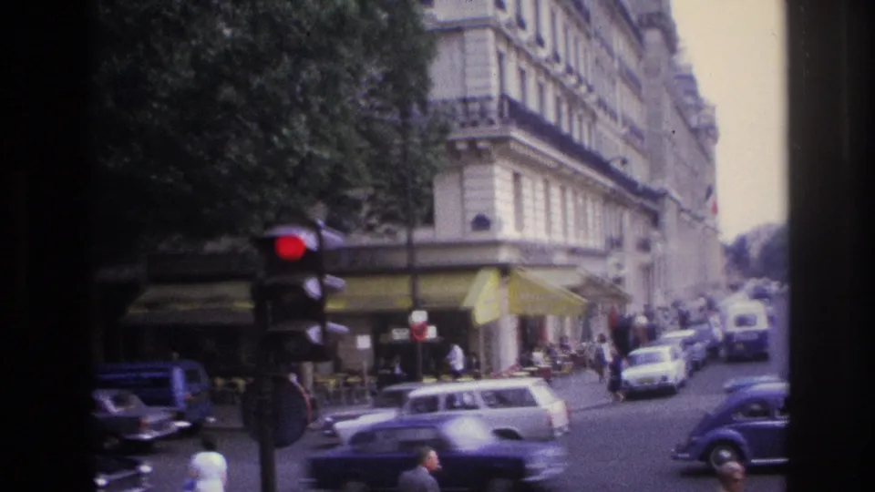 1967 Paris intersection with cars moving past a corner café, yellow awnings, pedestrians, and a red traffic signal.