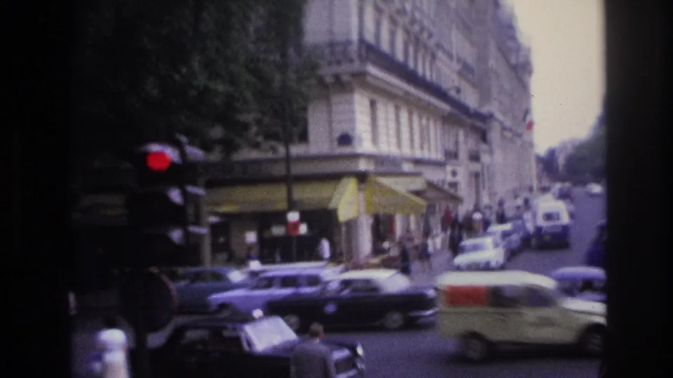 Handheld 1967 Paris street view showing a red traffic light, passing vintage cars, pedestrians, café awnings, and apartment façades.