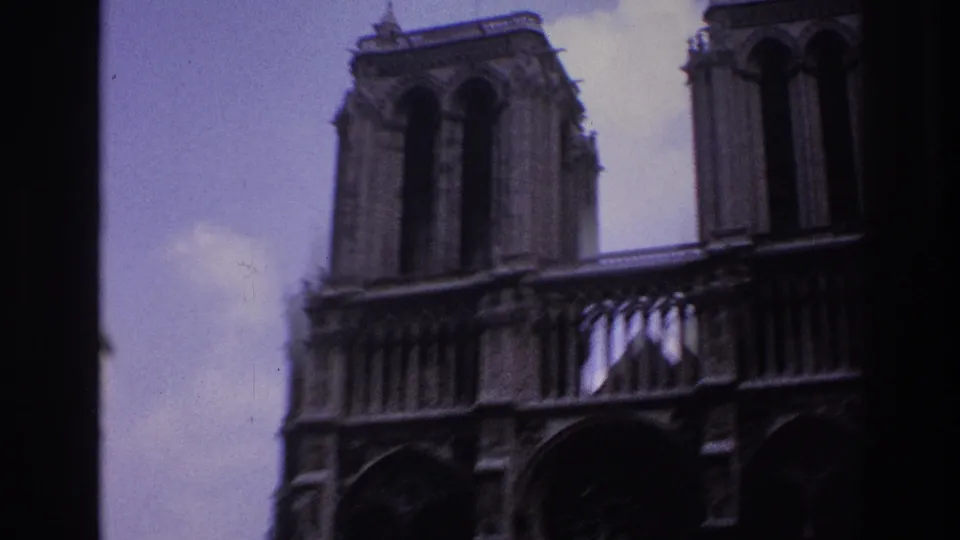 Soft 1967 Paris view of Notre-Dame's facade, partially framed by the tour-bus window edge and pale sky.