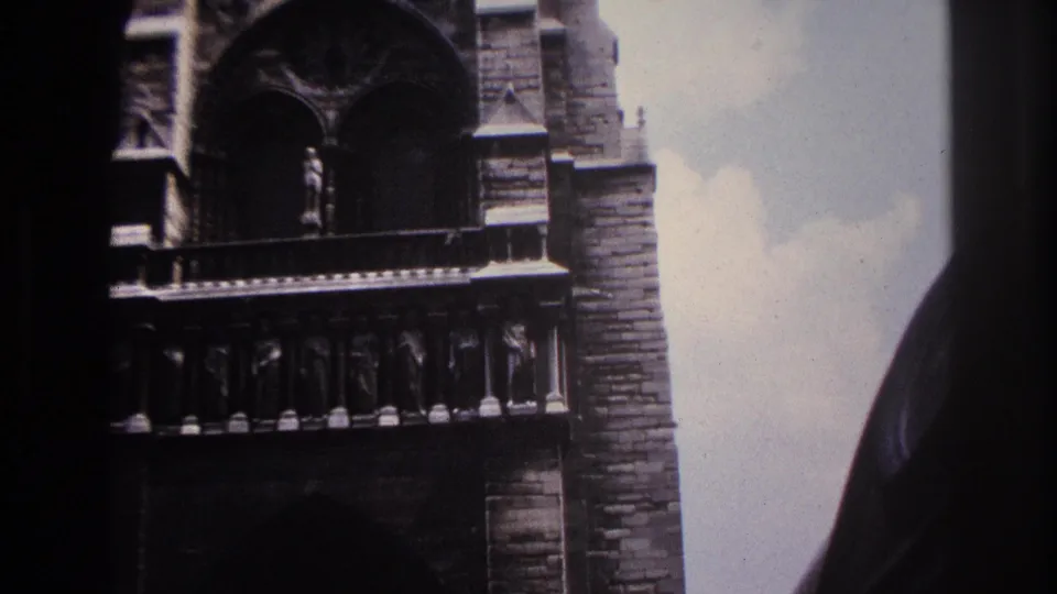 1967 Paris Super 8 frame showing a lower portion of a Gothic cathedral facade, with a dark arch, carved row, and pale sky at the side.