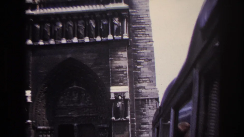 1967 Paris home-movie frame showing a Gothic cathedral facade partly obscured by the edge of a moving vehicle.