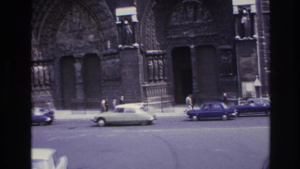 1967 Paris home-movie frame showing cars, pedestrians, railings, and a Gothic cathedral facade from a moving viewpoint.