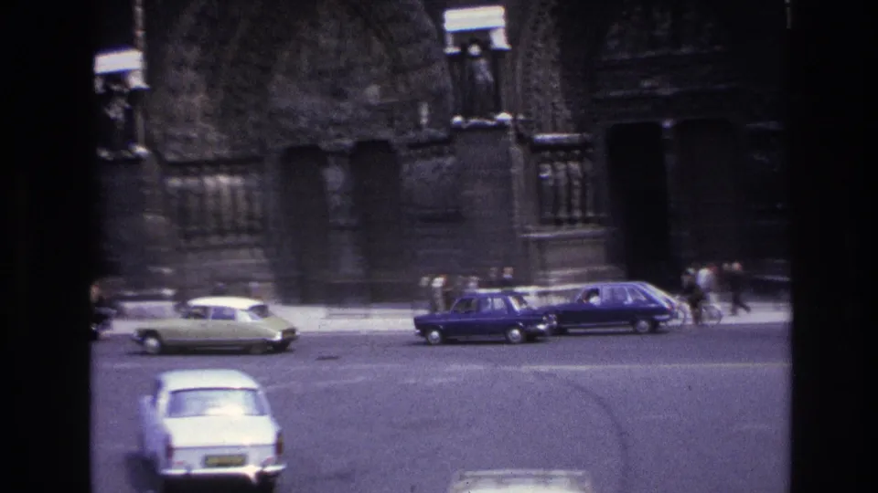 1967 Paris street view with small vintage cars crossing before a Gothic cathedral entrance and distant pedestrians.