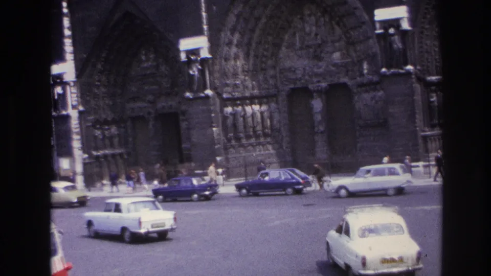 1967 Paris intersection with white and blue vintage cars moving past the ornate stone cathedral facade.