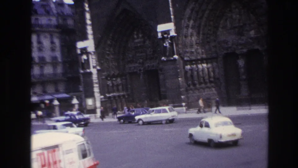 Paris 1967 home-movie frame with vintage traffic, a white car in the foreground, and the cathedral facade behind.