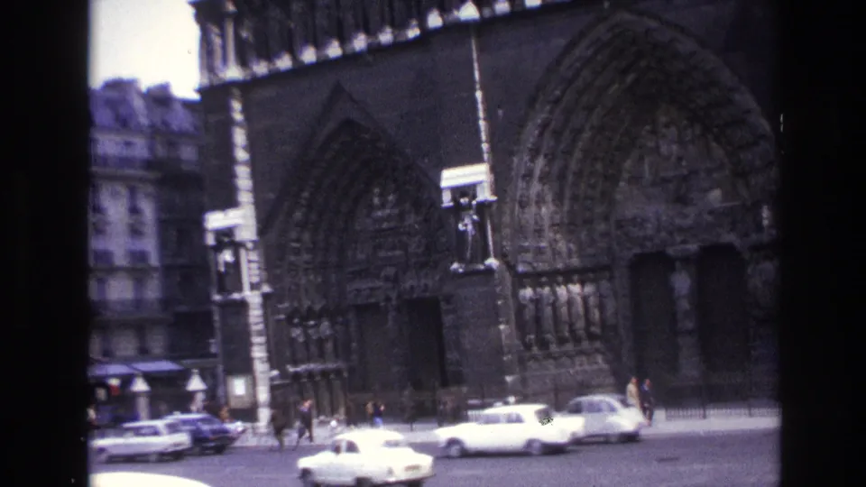 1967 Paris street scene with white vintage cars passing in front of a Gothic cathedral portal and sidewalk pedestrians.