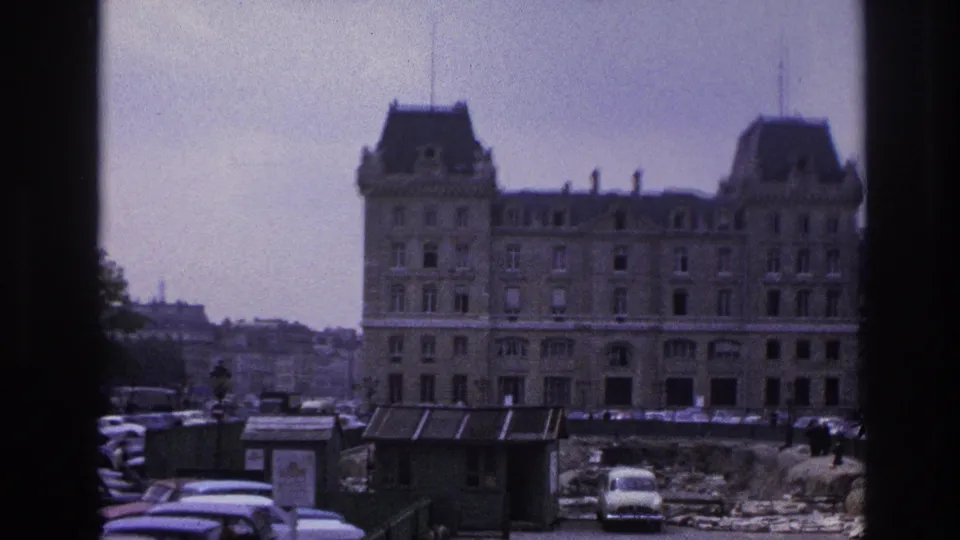 1967 Paris street frame showing a large historic building with mansard roofs, parked mid-century cars, construction ground, and foreground kiosks.
