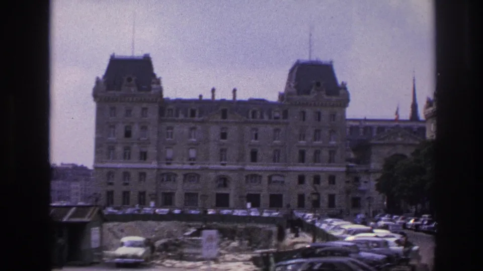 1967 Paris home-movie frame showing a wide street view with a large mansard-roofed building, parked cars, construction area, trees, and distant spires.