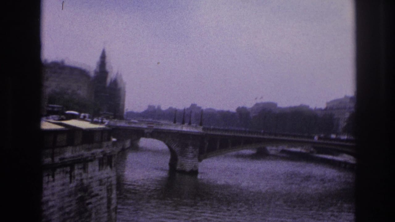 1967 Paris home-movie frame showing a river bridge, stone quay, distant spired tower, and soft purple-blue film tint.