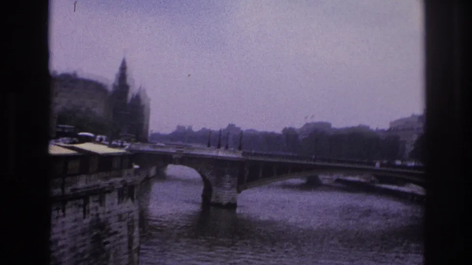 1967 Paris home-movie frame showing a river bridge, stone quay, distant spired tower, and soft purple-blue film tint.