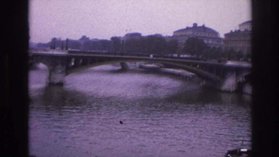 1967 Paris travel frame showing a broad river, arched bridge, tree line, and formal riverside buildings in soft focus.