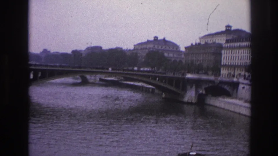 1967 Paris home-movie frame with bridge arches, rippled river water, quayside buildings, trees, and small distant street activity.