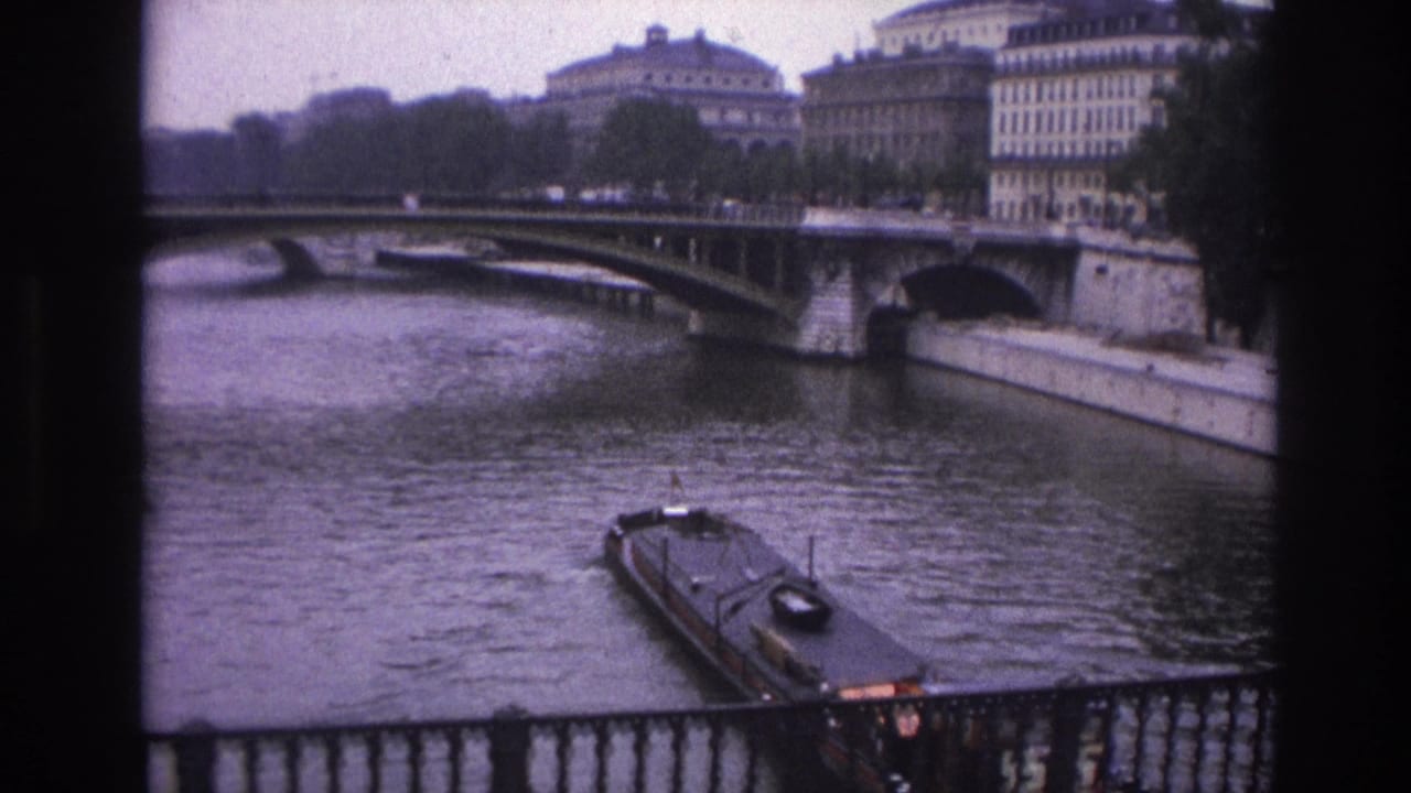 1967 Paris home-movie frame showing a barge moving on the Seine below a stone bridge, seen past a dark foreground border and railing.