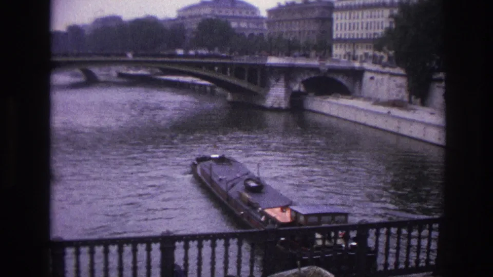 The barge has moved closer on the Seine, with a stone bridge, embankment, trees, and Paris buildings behind it.