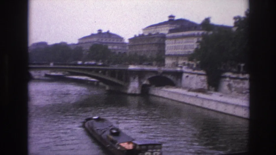Soft 8mm Paris view of a barge passing under or near a stone bridge on the Seine, with masonry banks and buildings in the background.
