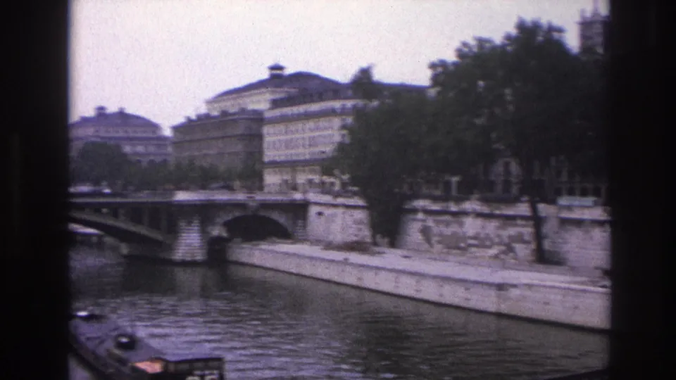 1967 Paris 8mm frame from the Seine showing a bridge, riverside buildings, trees, stone quay walls, and part of a barge.