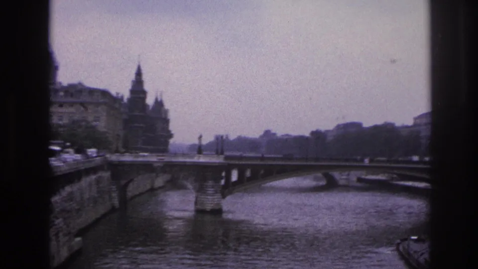 1967 Paris 8mm frame showing the Seine under a bridge, distant spired architecture, quay walls, parked cars, and part of a barge.