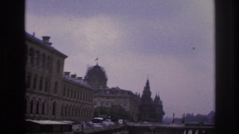 1967 Paris riverbank view with a large historic-looking building, bridge, distant spires, parked cars, and the Seine below.