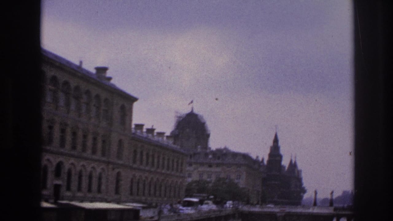 1967 Paris home-movie frame showing ornate stone buildings, distant pedestrians, and street traffic through dark vertical framing.