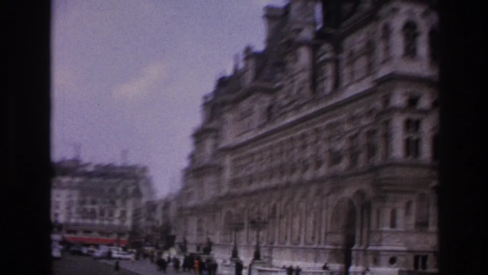 1967 Paris street view with a large historic facade, rows of windows, pedestrians, and vehicles in soft-focus 8mm color.