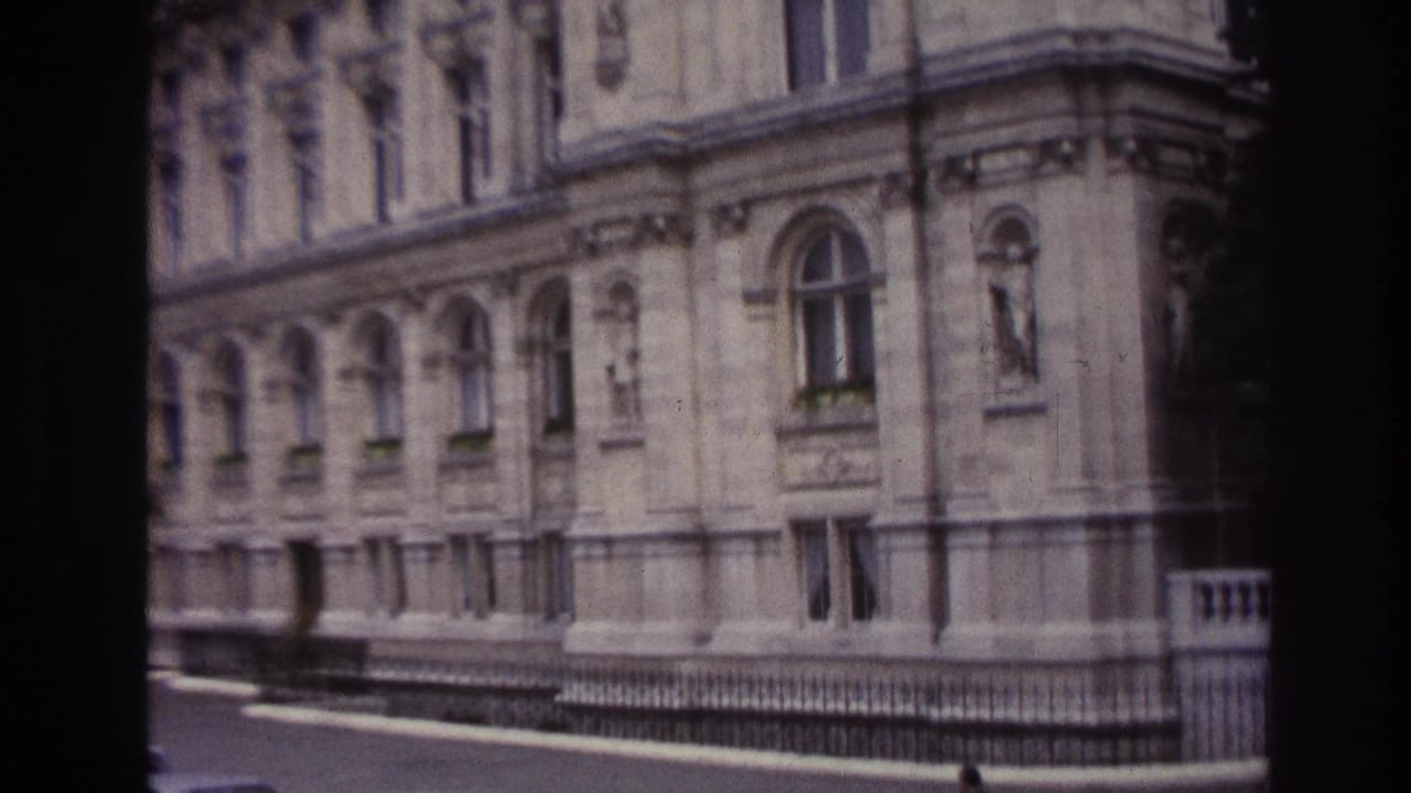 1967 Paris home-movie frame showing a large classical stone facade, arched windows, railings, and a quiet street edge.
