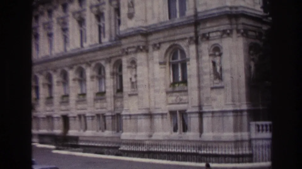 1967 Paris home-movie frame showing a large classical stone facade, arched windows, railings, and a quiet street edge.