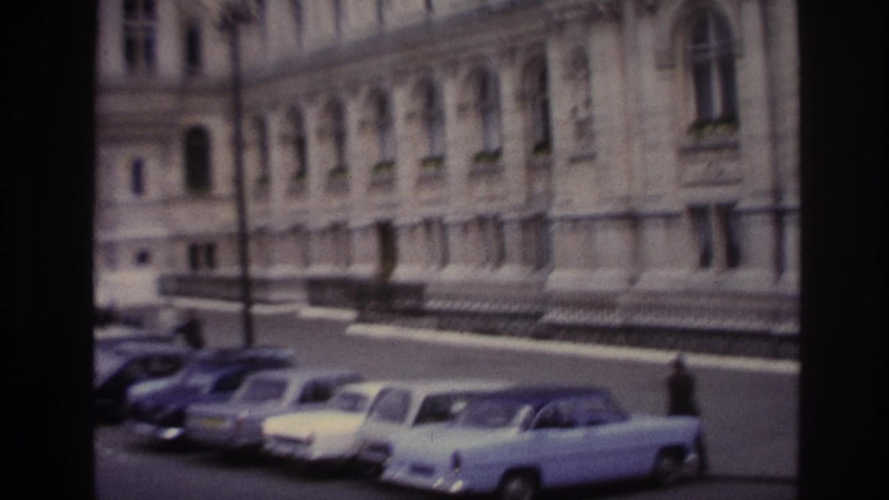 Parked 1960s cars line the road beside a large ornate stone building.