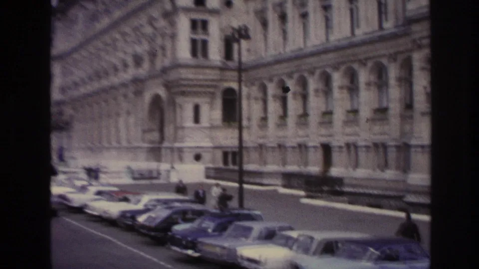 1967 Paris home-movie frame showing a row of parked cars and small pedestrians outside a large classical building.