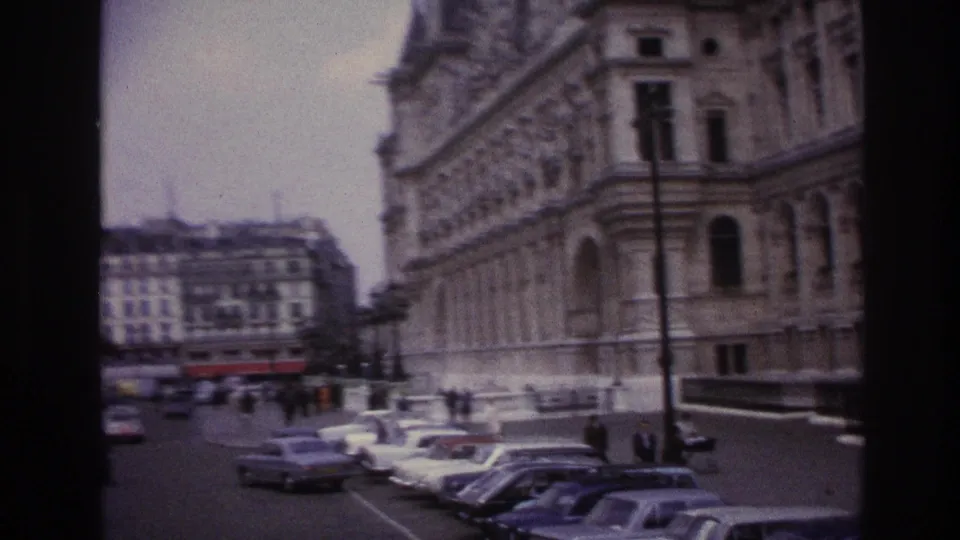 Wide 1967 Paris street frame with parked cars, pedestrians, lampposts, and the corner of a classical stone building.
