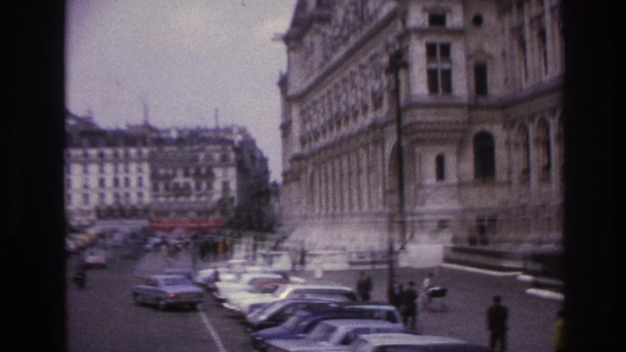 1967 Paris street view with parked cars, small pedestrians, and historic building facades seen in soft 8mm color.