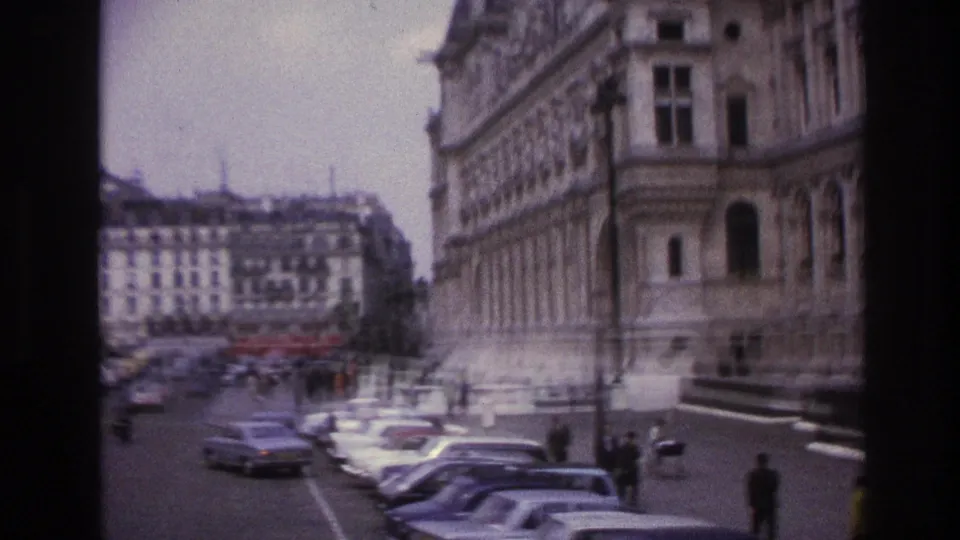 1967 Paris street view with parked cars, small pedestrians, and historic building facades seen in soft 8mm color.