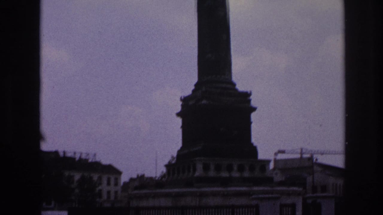 1967 Paris home-movie frame showing a monument base through dark window edges with mid-century cars moving across the street.
